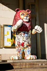 Eine farbenfroh gestaltete Skulptur eines aufrecht stehenden Löwen ziert den Eingangsbereich der Leipziger Oper am Augustusplatz. Das Gehäuse der kunstvollen Löwenfigur ist über und über mit detailreichen, bunten Motiven bemalt, welche an klassische Opernszenen und historische Kostüme erinnern. Als symbolisches Wappentier der Stadt Leipzig schlägt diese kreative Kunstinstallation im öffentlichen Raum eine charmante Brücke zwischen dem traditionellen Stadtwappen und dem modernen kulturellen Leben vor dem Theatergebäude.