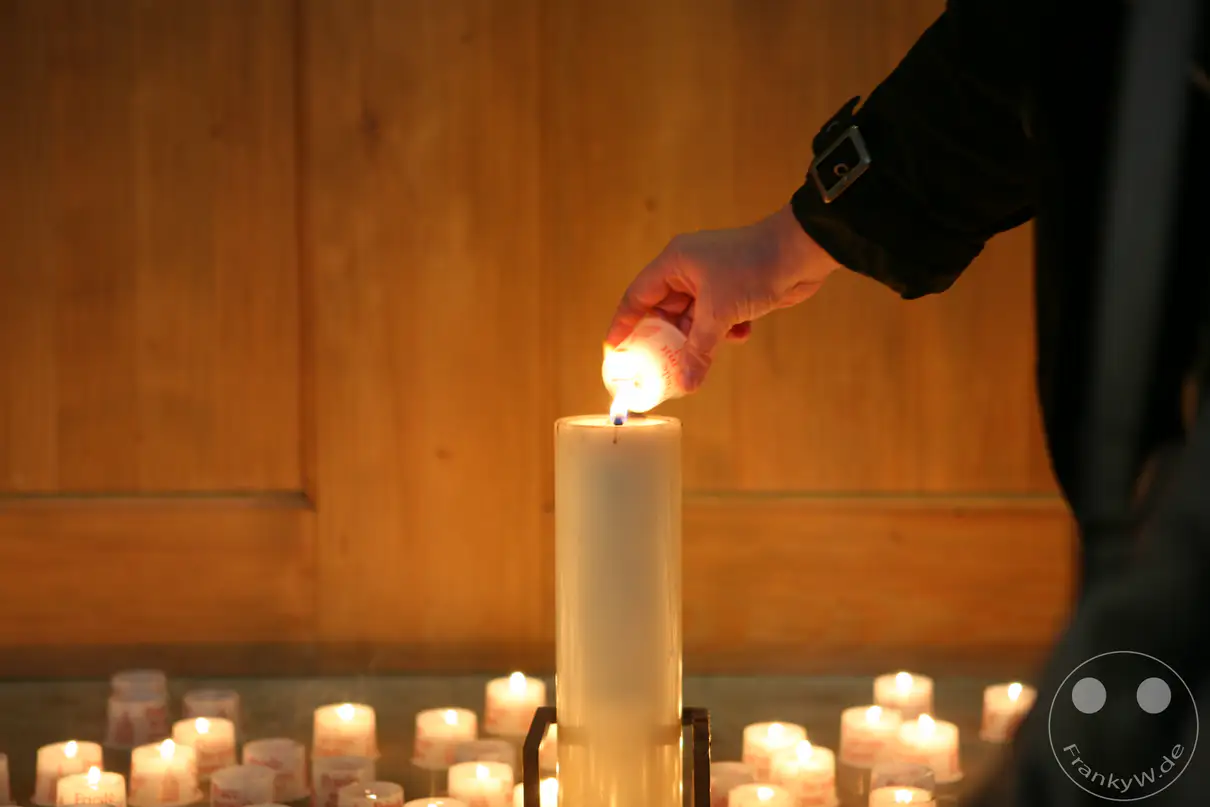 In der Frauenkirche in Dresden wird eine Kerze angezündet, während viele weitere Kerzen vor einer Holzvertäfelung brennen. Das gedämpfte Licht und die Flammen erzeugen eine besinnliche und feierliche Atmosphäre. Diese Szene vermittelt ein Gefühl von Frieden und gemeinsamer Erinnerung in der historischen Kirche.
