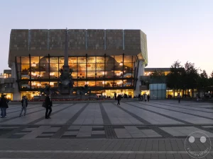 Der Augustusplatz in Leipzig erstrahlt im Abendlicht vor dem imposanten Gewandhaus. Die großflächige Platzgestaltung aus dunklen und hellen Steinen bildet einen modernen Kontrast zur beleuchteten Fassade des Konzerthauses. Passanten bewegen sich über den weitläufigen Platz, während der Himmel in sanften Pastelltönen ausklingt.