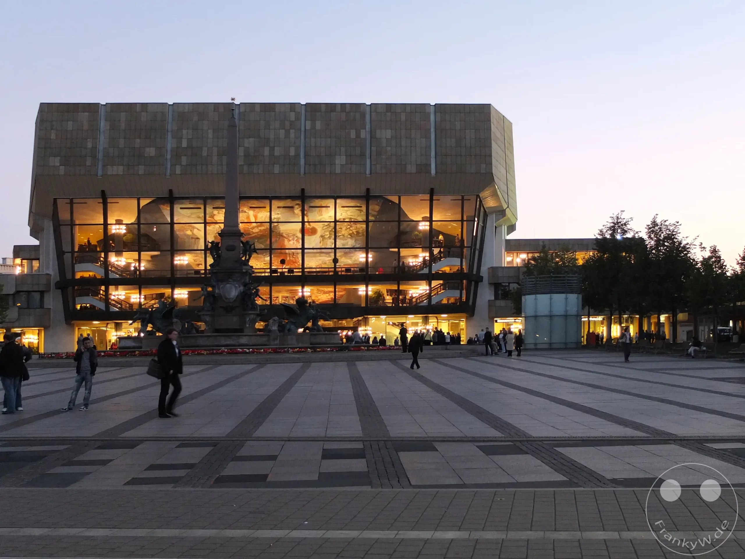 Der Augustusplatz in Leipzig erstrahlt im Abendlicht vor dem imposanten Gewandhaus. Die großflächige Platzgestaltung aus dunklen und hellen Steinen bildet einen modernen Kontrast zur beleuchteten Fassade des Konzerthauses. Passanten bewegen sich über den weitläufigen Platz, während der Himmel in sanften Pastelltönen ausklingt.