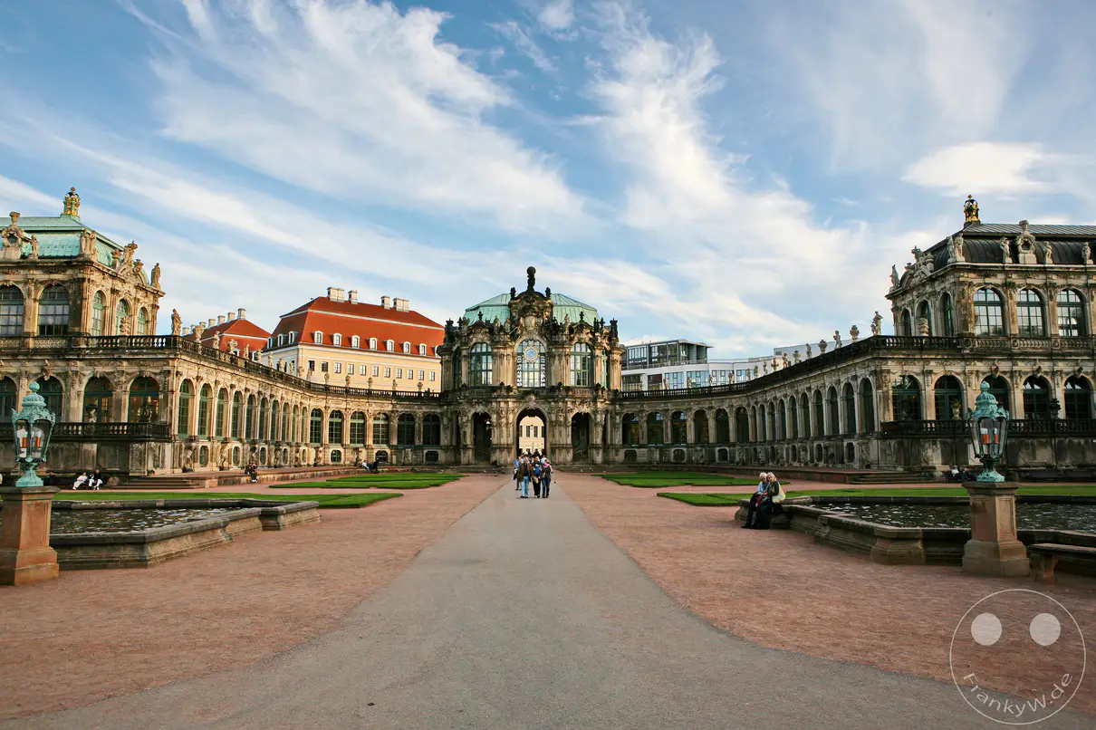 Der Dresdner Zwinger in Deutschland, eine prächtige barocke Schlossanlage, präsentiert sich mit symmetrischen Arkaden, kunstvollen Skulpturen und grünen Rasenflächen. Besucher schlendern durch den weitläufigen Innenhof, während die Sonne auf die beeindruckende Architektur fällt und eine friedliche Atmosphäre schafft. Die Szene ist geprägt von der Pracht des Barockstils und lädt zum Verweilen ein.