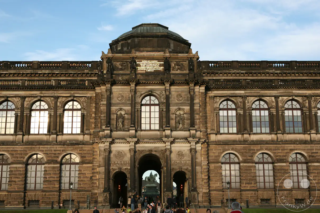 Die imposante Fassade des Dresdner Zwingers, ein Meisterwerk barocker Architektur, erhebt sich unter einem blauen Himmel mit wenigen Wolken. Menschenscharen bewegen sich vor dem Haupteingang, einige bestaunen die Skulpturen, andere treten ein, um die reichen Sammlungen zu erkunden. Die warme Nachmittagssonne wirft Glanzlichter auf die Fenster und verleiht der Szene eine lebendige und geschichtsträchtige Atmosphäre.