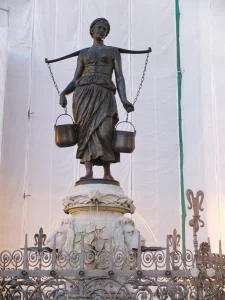 Die Bronzefigur einer Wasserträgerin ist zentral auf dem historischen Mägdebrunnen in Leipzig positioniert. Mit zwei Eimern an einer Stange stellt die Skulptur das frühere Leben und die Wasserversorgung der Stadt dar. Eine atmosphärische Darstellung von Handwerk und Stadtgeschichte, eingefangen vor einem weiß verhüllten Hintergrund, der auf laufende Arbeiten hindeutet.