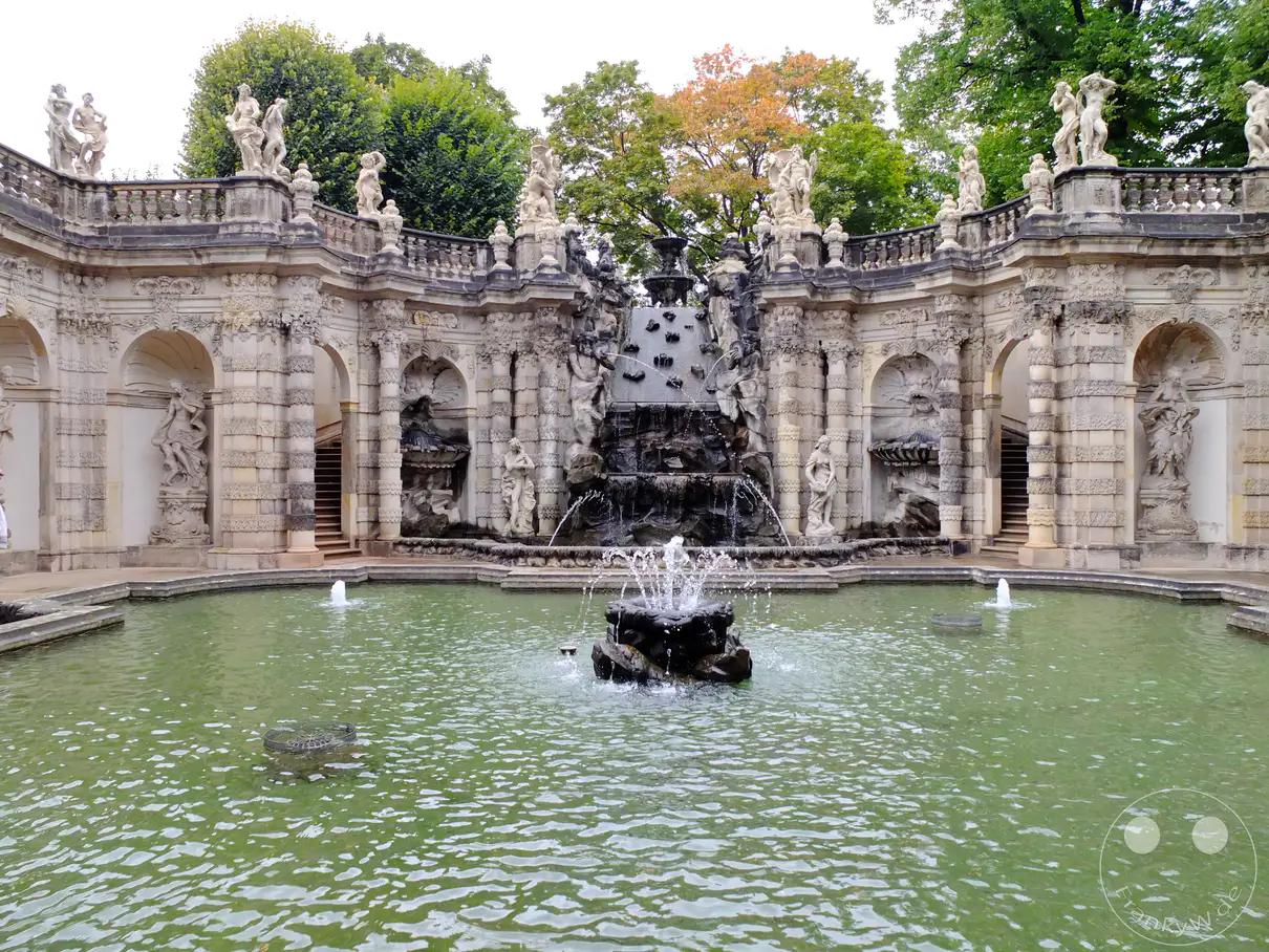 Das Nymphenbad im Dresdner Zwinger ist ein Meisterwerk barocker Gartenkunst. Eine kunstvolle Fontäne sprudelt in einem grünen Wasserbecken, umgeben von beeindruckenden Skulpturen und architektonischen Elementen. Die Szene strahlt Ruhe und Erhabenheit aus und lädt zum Verweilen ein.