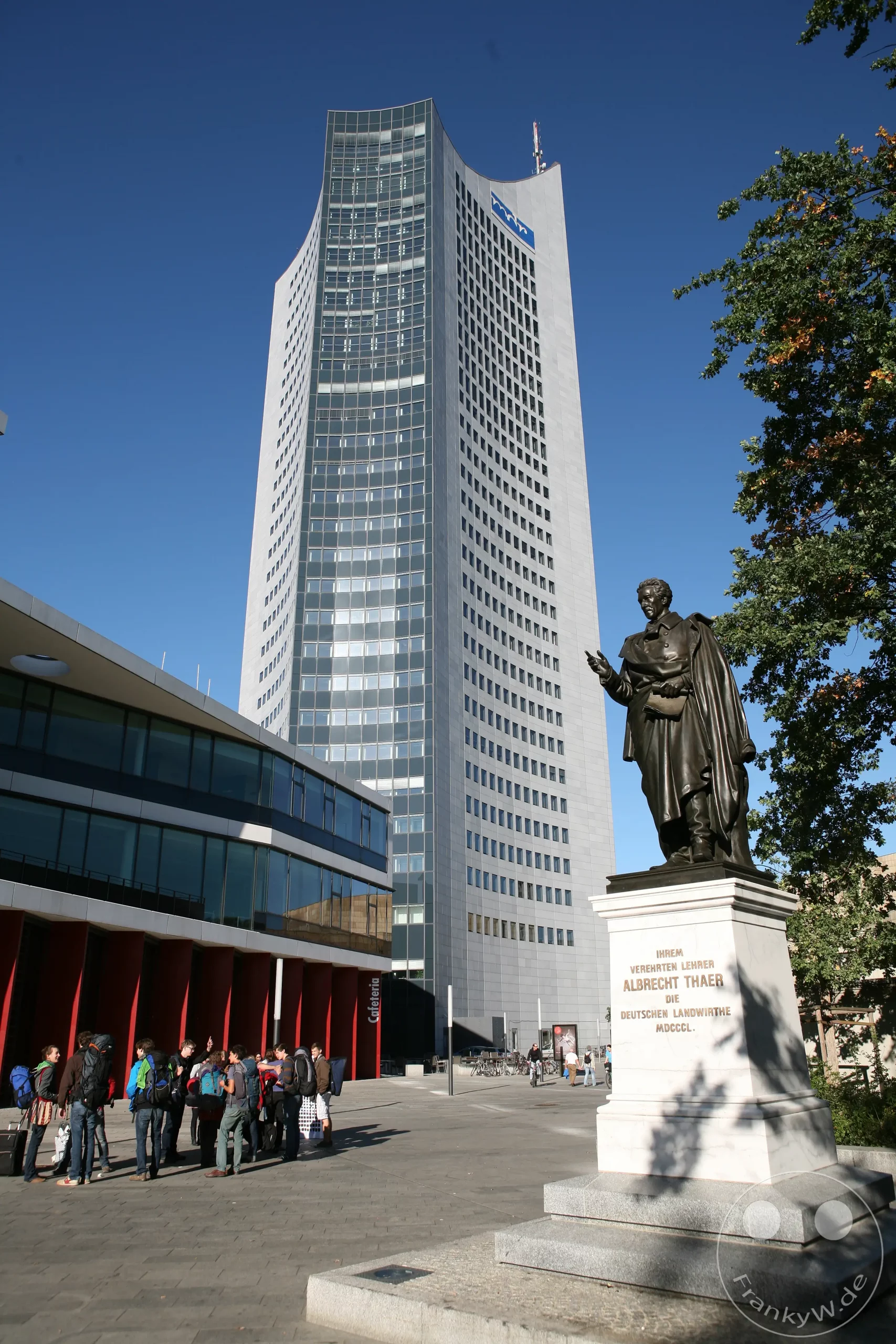 Das Albrecht-Thaer-Denkmal steht vor einem modernen Bürogebäude, wahrscheinlich dem MDR-Gebäude, in Leipzig, Deutschland. Eine Gruppe von Menschen mit Rucksäcken versammelt sich auf einem öffentlichen Platz, während die Sonne auf die klare blaue Himmelskulisse scheint. Die Szene vermittelt eine Atmosphäre von Urbanität und Bildung.