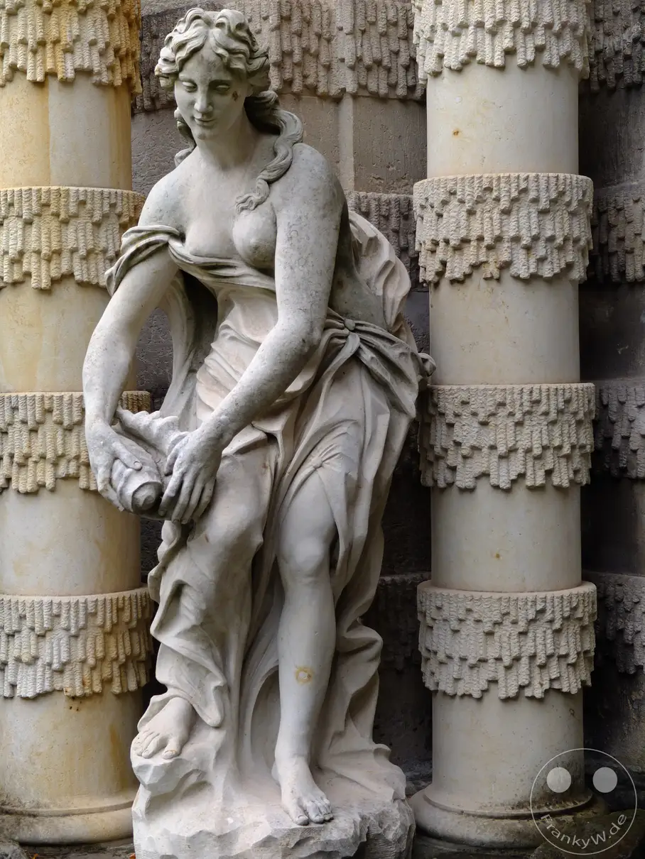 Eine kunstvolle Steinskulptur einer Nymphe, die in einem opulenten Nymphenbad im Zwinger in Dresden ruht. Die feinen Details der Statue und die umgebende Architektur vermitteln ein Gefühl von historischer Pracht und stiller Anmut. Die Wassergöttin scheint in ihrem Element, umgeben von den kunstvollen Verzierungen des barocken Bades.
