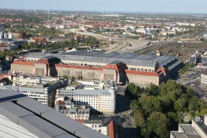 Blick aus der Vogelperspektive auf Leipzig zeigt die beeindruckende Architektur des Hauptbahnhofs, flankiert von zahlreichen Gleisanlagen und urbanen Gebäuden. Umgeben von Grünflächen und einer weiten Stadtsilhouette unter einem strahlend blauen Himmel, vermittelt die Szene einen Eindruck von der Größe und Betriebsamkeit der sächsischen Metropole. Die Komposition aus historischen und modernen Elementen spiegelt die dynamische Entwicklung der Stadt wider.