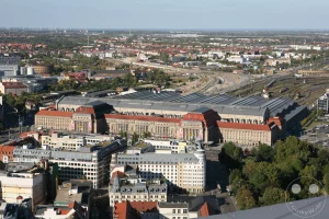 Die beeindruckende Architektur des Leipziger Hauptbahnhofs wird aus der Vogelperspektive präsentiert. Zahlreiche Bahngleise und ein weitläufiges Stadtbild von Leipzig erstrecken sich in der Ferne unter einem klaren Himmel. Die Aufnahme vermittelt einen Eindruck von der urbanen Weite und der Bedeutung des Eisenbahnknotenpunkts.