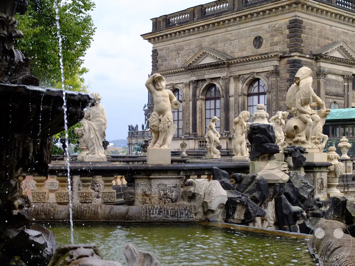 Das obere Wasserbecken des Nymphenbads im Dresdner Zwinger ist mit kunstvollen Skulpturen geschmückt, darunter ein Satyr (Faun), der auf einem Meereswesen reitet. Im Hintergrund erhebt sich der Französische Pavillon mit seiner beeindruckenden Architektur. Die Szenerie strahlt eine königliche Eleganz und historische Bedeutung aus.