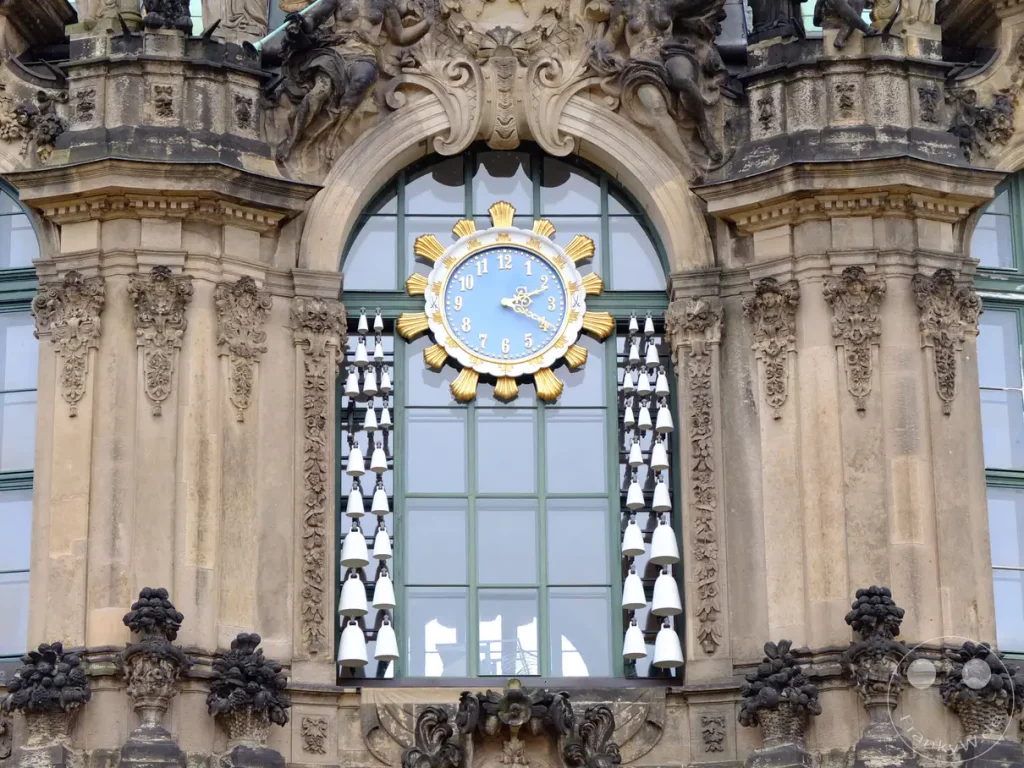 Das Meissener Porzellan-Glockenspiel im Zwinger in Dresden erklingt mit seinem charmanten Klang. Zahlreiche weiße Glocken hängen in Reih und Glied vor dem großen Fenster und dem prächtigen Uhrwerk. Die barocke Fassade mit ihren aufwendigen Skulpturen und Verzierungen strahlt eine würdevolle Eleganz aus.