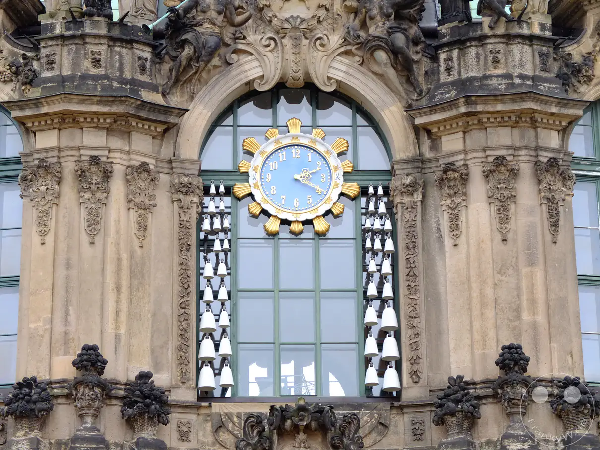 Das Meissener Porzellan-Glockenspiel im Zwinger in Dresden erklingt mit seinem charmanten Klang. Zahlreiche weiße Glocken hängen in Reih und Glied vor dem großen Fenster und dem prächtigen Uhrwerk. Die barocke Fassade mit ihren aufwendigen Skulpturen und Verzierungen strahlt eine würdevolle Eleganz aus.