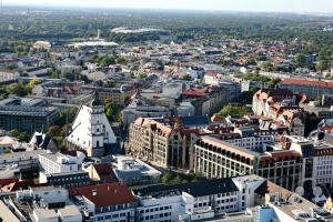 Die weitläufige Luftaufnahme überblickt das dicht bebaute Leipziger Stadtzentrum mit seinen charakteristischen roten Ziegeldächern und historischen Bauten. Als markanter Orientierungspunkt sticht die geschichtsträchtige Thomaskirche mit ihrem hohen, spitzen Kirchendach und dem weißen Turm aus der urbanen Struktur hervor. Am rechten Bildrand zeigt sich ein Teil des Alten Rathauses am Markt, während sich im Hintergrund die grünen Ausläufer des Rosentals und das markante Rund der Red Bull Arena am Horizont abzeichnen.