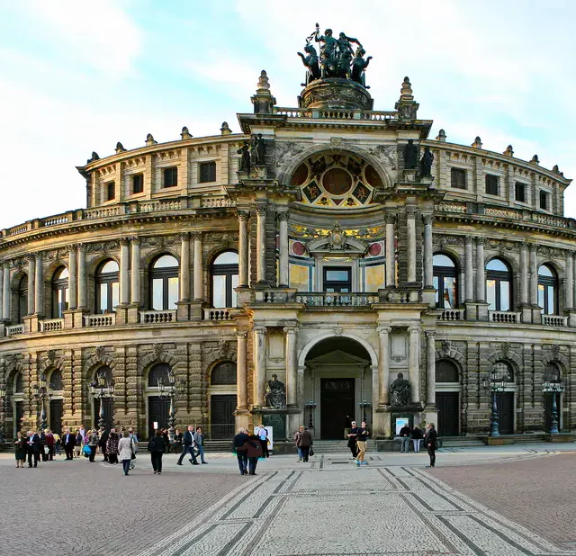 Die prunkvolle Semperoper in Dresden, ein Meisterwerk der Architektur, erhebt sich vor einem weiten Platz, auf dem sich Besucher bewegen. Die Fassade des Opernhauses beeindruckt mit zahlreichen Verzierungen und Skulpturen, umgeben von einem blauen Himmel mit vereinzelten Wolken. Die Szene strahlt eine würdevolle und kulturelle Atmosphäre aus, die die historische Bedeutung des Ortes unterstreicht.