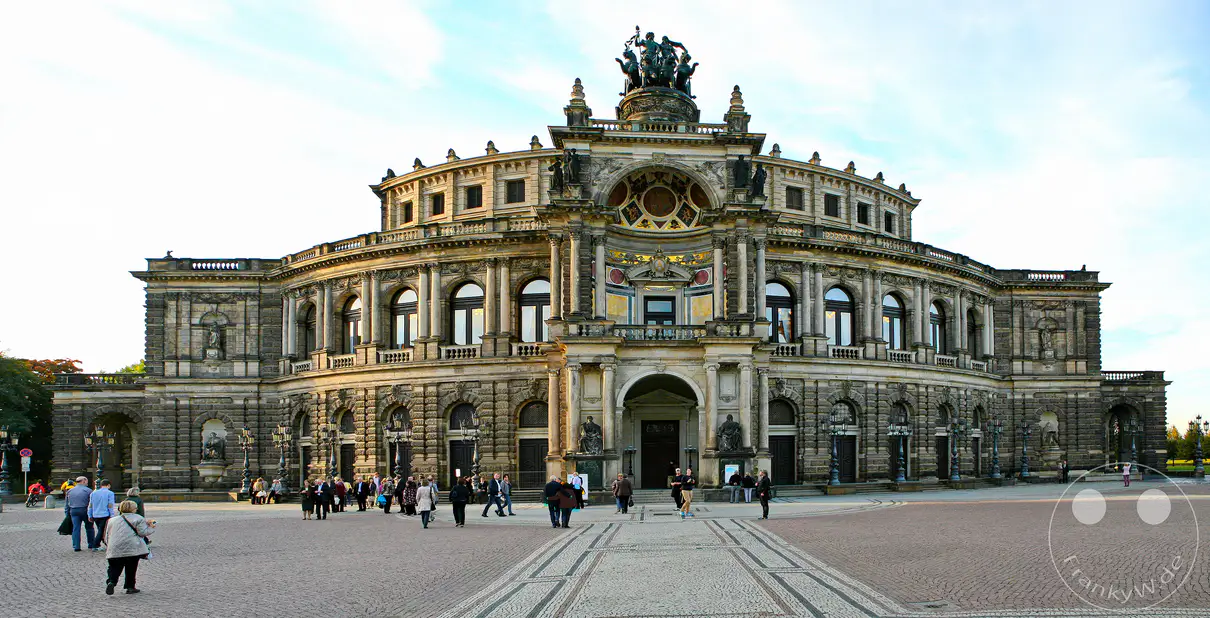 Die prunkvolle Semperoper in Dresden, ein Meisterwerk der Architektur, erhebt sich vor einem weiten Platz, auf dem sich Besucher bewegen. Die Fassade des Opernhauses beeindruckt mit zahlreichen Verzierungen und Skulpturen, umgeben von einem blauen Himmel mit vereinzelten Wolken. Die Szene strahlt eine würdevolle und kulturelle Atmosphäre aus, die die historische Bedeutung des Ortes unterstreicht.