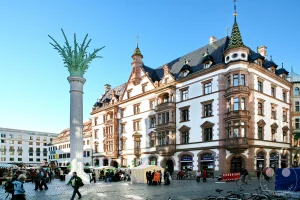 Ein lebhafter Marktplatz in Leipzig, Deutschland, wird von der historischen Nikolaisäule und dem markanten Predigerhaus dominiert. Menschen flanieren zwischen Marktständen und bestaunen die Detailreiche Architektur der Gebäude, während ein klarer Himmel die Szenerie überstrahlt. Die Atmosphäre ist friedlich und geschäftig zugleich, typisch für einen sonnigen Tag in einer europäischen Stadt.