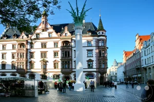 Ein lebhafter Marktplatz in Leipzig, Deutschland, wird von der historischen Nikolaisäule und dem markanten Predigerhaus dominiert. Menschen flanieren zwischen Marktständen und bestaunen die Detailreiche Architektur der Gebäude, während ein klarer Himmel die Szenerie überstrahlt. Die Atmosphäre ist friedlich und geschäftig zugleich, typisch für einen sonnigen Tag in einer europäischen Stadt.