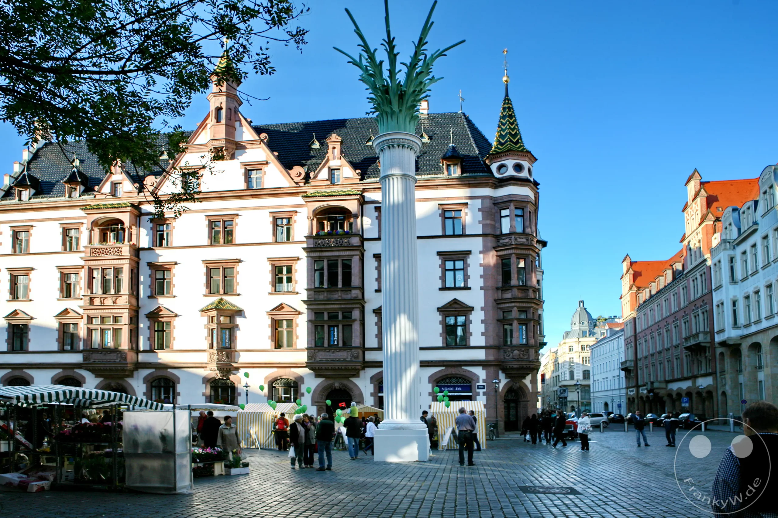 Ein lebhafter Marktplatz in Leipzig, Deutschland, wird von der historischen Nikolaisäule und dem markanten Predigerhaus dominiert. Menschen flanieren zwischen Marktständen und bestaunen die Detailreiche Architektur der Gebäude, während ein klarer Himmel die Szenerie überstrahlt. Die Atmosphäre ist friedlich und geschäftig zugleich, typisch für einen sonnigen Tag in einer europäischen Stadt.