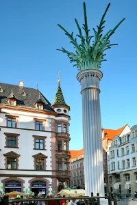 Ein lebhafter Marktplatz in Leipzig, Deutschland, wird von der historischen Nikolaisäule und dem markanten Predigerhaus dominiert. Menschen flanieren zwischen Marktständen und bestaunen die Detailreiche Architektur der Gebäude, während ein klarer Himmel die Szenerie überstrahlt. Die Atmosphäre ist friedlich und geschäftig zugleich, typisch für einen sonnigen Tag in einer europäischen Stadt.