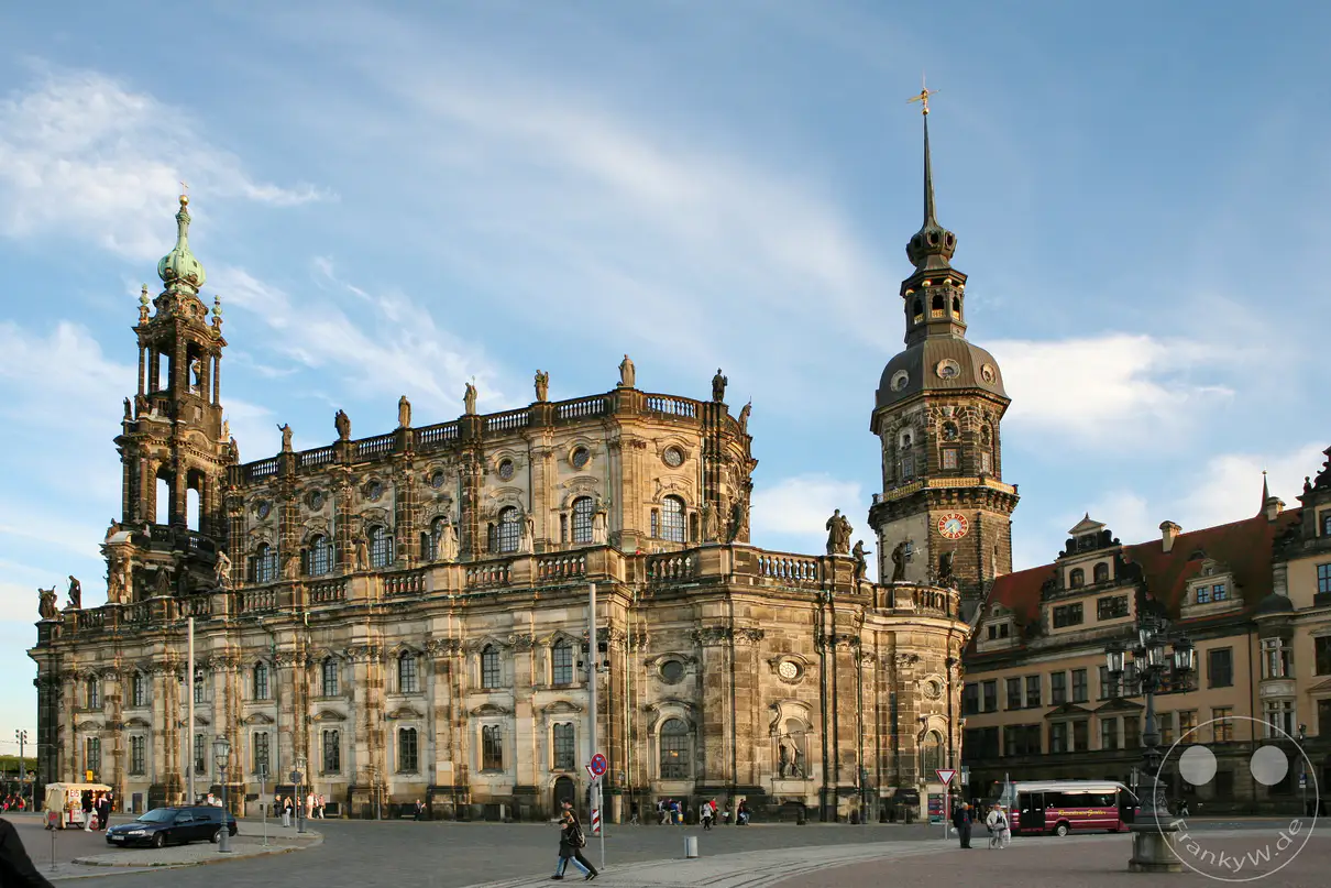 Die barocke Katholische Hofkirche erstreckt sich hier in ihrer vollen Längsansicht im Herzen der Dresdner Altstadt. Das monumentale Gotteshaus, das heute als Kathedrale Sanctissimae Trinitatis dient, besticht durch seine reich verzierte Fassade und die charakteristischen Heiligenstatuen auf den Balustraden. Rechts im Hintergrund ragt der Hausmannsturm des angrenzenden Residenzschlosses empor, dessen Architektur fließend in die Renaissancebauten des ehemaligen Herrschersitzes übergeht. Zusammen bilden diese beiden historischen Bauwerke eines der bekanntesten und am häufigsten fotografierten Ensembles der sächsischen Landeshauptstadt.