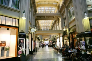 Eine elegante Einkaufspassage in Leipzig, Deutschland, mit kunstvoller Architektur und einer gläsernen Decke, die das Innere in ein warmes Licht taucht. Menschen flanieren zwischen den Geschäften und Cafés, deren Schilder auf die Vielfalt des Angebots hinweisen. Die Szene strahlt eine Atmosphäre von urbanem Leben und historischem Charme aus.