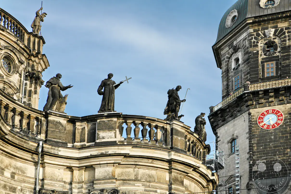 Mehrere monumentale Heiligenstatuen aus Sandstein thronen auf der geschwungenen Balustrade der barocken Katholischen Hofkirche. Diese von Lorenzo Mattielli geschaffenen Skulpturen prägen die Dachlandschaft der Kathedrale und stellen wichtige Heilige sowie Ordensgründer dar. Im rechten Hintergrund ragt die dunkle Fassade des Hausmannsturms vom Dresdner Residenzschloss empor, dessen goldene Turmuhr mit den römischen Ziffern deutlich zu erkennen ist. Das Zusammenspiel aus den sakralen Figuren und dem herrschaftlichen Turm verdeutlicht die enge architektonische und historische Verbindung der beiden Baudenkmäler.
