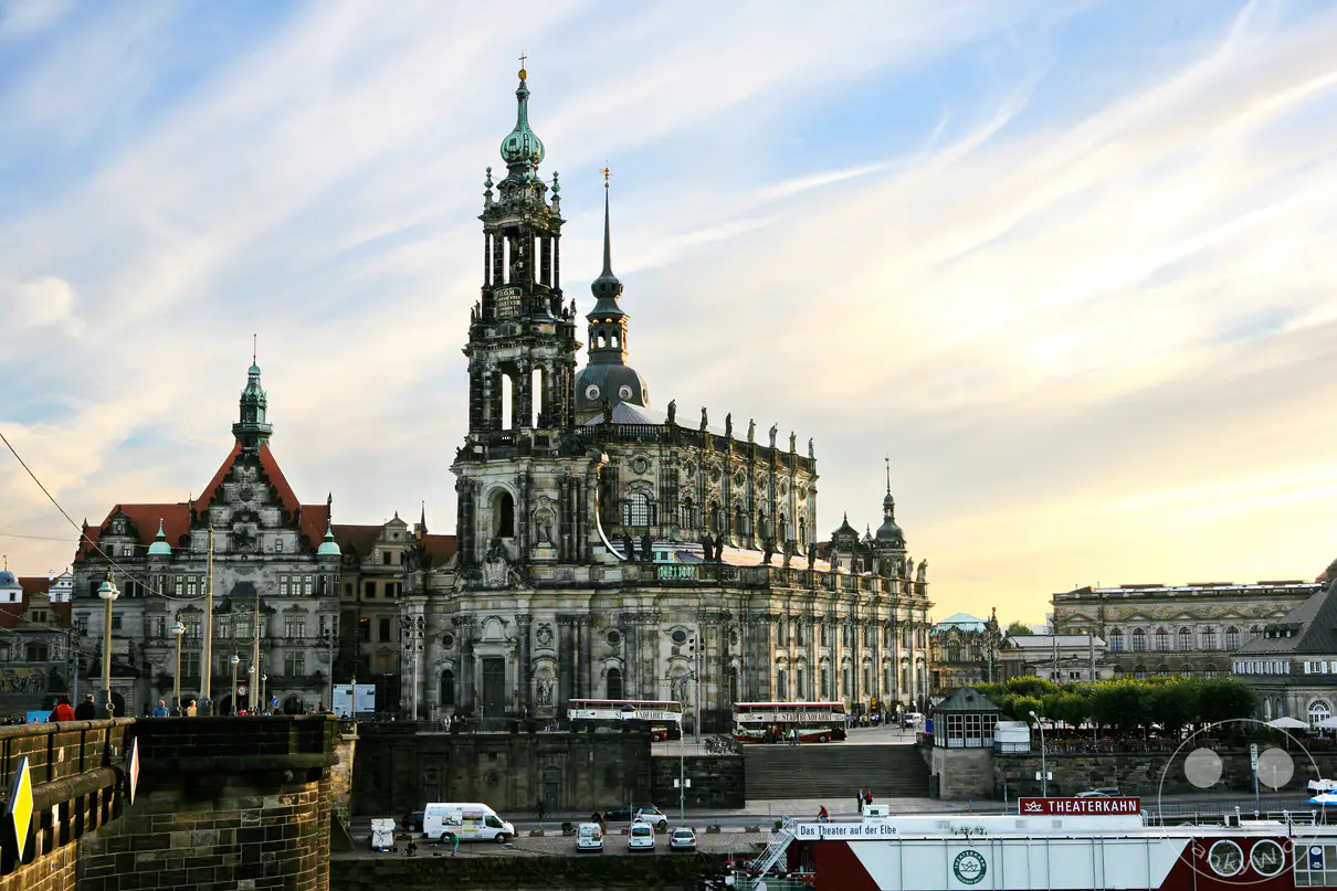 Die imposante Kathedrale Sanctissimae Trinitatis thront majestätisch über dem Elbufer in Dresden. Prächtige Architektur und historische Gebäude prägen das Stadtbild unter einem weitläufigen Himmel mit leichten Wolken. Die Abendsonne taucht die Szenerie in ein warmes Licht und schafft eine friedliche Atmosphäre.