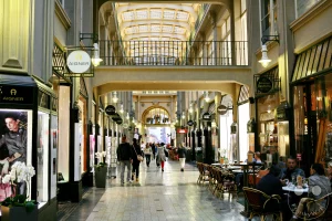 Eine elegante Einkaufspassage in Leipzig, Deutschland, mit kunstvoller Architektur und einer gläsernen Decke, die das Innere in ein warmes Licht taucht. Menschen flanieren zwischen den Geschäften und Cafés, deren Schilder auf die Vielfalt des Angebots hinweisen. Die Szene strahlt eine Atmosphäre von urbanem Leben und historischem Charme aus.