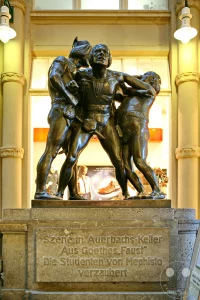 Die Bronzeplastik "Szene in Auerbachs Keller" nach Goethes Faust von Carl Seffner steht in Leipzig. Dargestellt ist die Figurengruppe, die die legendäre Szene "Die Studenten von Mephisto verzaubert" zum Leben erweckt, ein ikonisches Werk im Kontext der deutschen Literaturgeschichte. Die Darstellung strahlt eine dynamische und theatralische Atmosphäre aus, eingefangen vor dem Hintergrund eines Schaufensters.