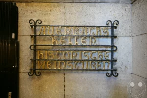 Das kunstvoll geschmiedete historische Metallgitter an einer hellen Sandsteinwand markiert den Eingangsbereich zu Auerbachs Keller in der Mädlerpassage. Die verzierten, goldglänzenden Jugendstil-Lettern kündigen die traditionsreichen historischen Weinstuben an, die zu den weltweit bekanntesten Gaststätten gehören und durch Goethes Drama Faust Weltruhm erlangten. Das dekorative Schmiedeeisen fügt sich nahtlos in die prachtvolle Architektur des historischen Messehauses ein und weist Einheimischen wie Touristen den Weg in das geschichtsträchtige Kellergewölbe.