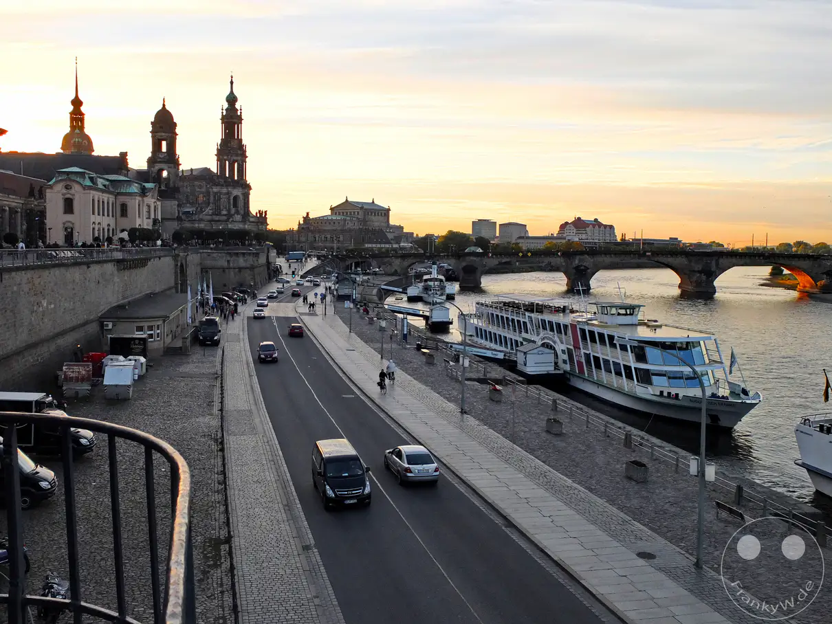 Ein malerisches Panorama von Dresden an der Elbe bei Sonnenuntergang, mit Blick auf die historische Altstadt und bedeutende Bauwerke wie die Hofkirche und die Semperoper. Schiffe der S�chsischen Dampfschifffahrt liegen am Terrassenufer vor Anker, w�hrend Autos und Radfahrer die Uferpromenade beleben. Die untergehende Sonne taucht den Himmel in warme Farben und schafft eine friedliche Abendstimmung �ber der s�chsischen Hauptstadt.