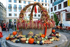 Eine beeindruckende Herbstdekoration mit Früchten, Gemüse und Blumen schmückt den Erntedankbrunnen auf dem Nikolaikirchhof in Leipzig, Deutschland. Die üppige Anordnung aus Kürbissen, Trauben, Äpfeln und Blumen strahlt eine festliche Erntestimmung aus. Im Hintergrund sind Gebäude und Passanten zu sehen, die das städtische Ambiente des Marktes unterstreichen.