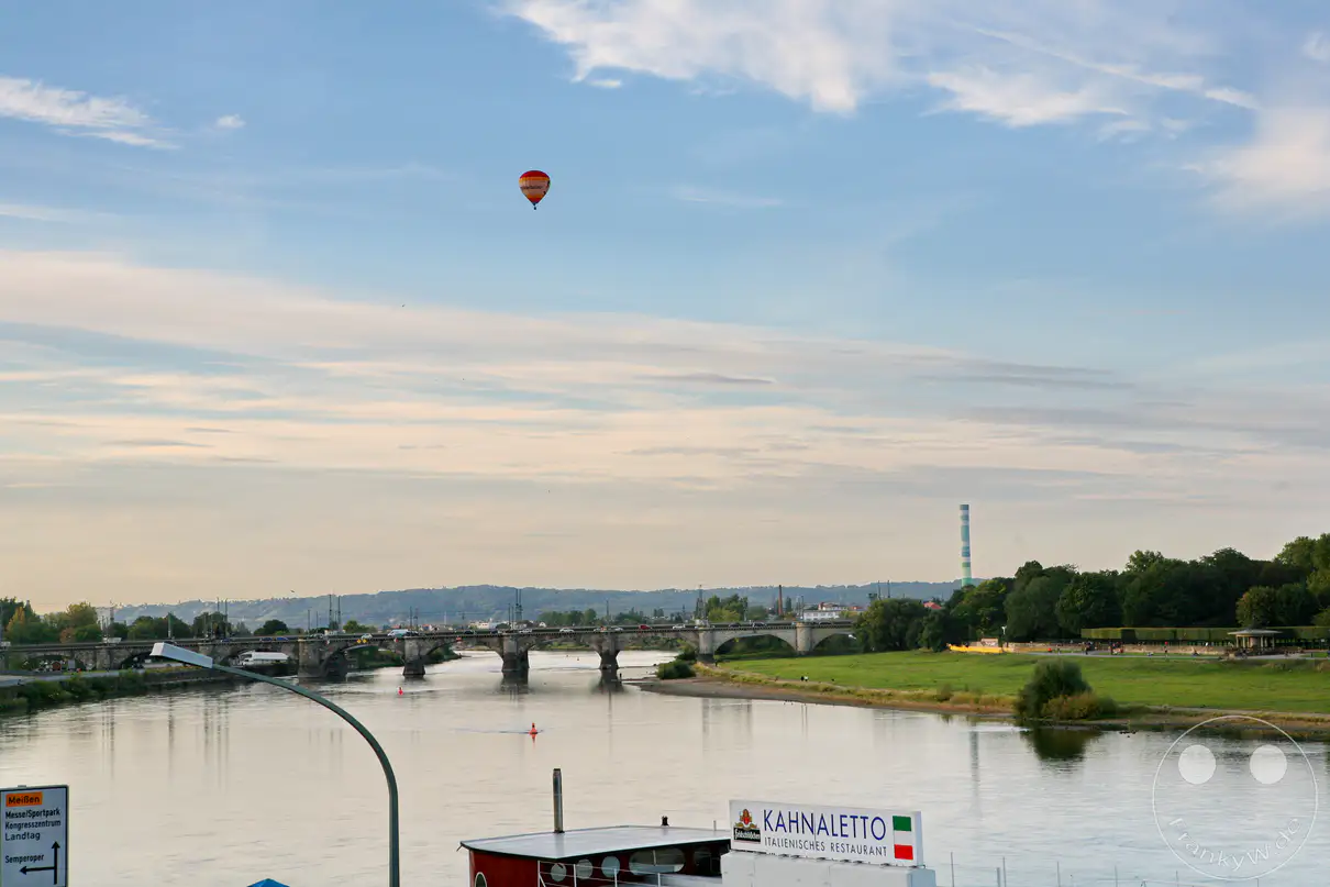Die Elbe schlängelt sich durch die Dresdner Landschaft, während im Hintergrund die historische Marienbrücke den Fluss überspannt. Am rechten Flussufer erstrecken sich die weitläufigen Elbwiesen, hinter denen ein gestreifter Industrieschornstein in den Himmel ragt. Ein farbiger Heißluftballon schwebt hoch über der Szenerie unter einem leicht bewölkten Abendhimmel. Im Vordergrund liegt das schwimmende italienische Restaurant „Kahnaletto“ am Ufer vertäut, während am linken Bildrand ein Straßenschild den Weg zum Kongresszentrum weist.