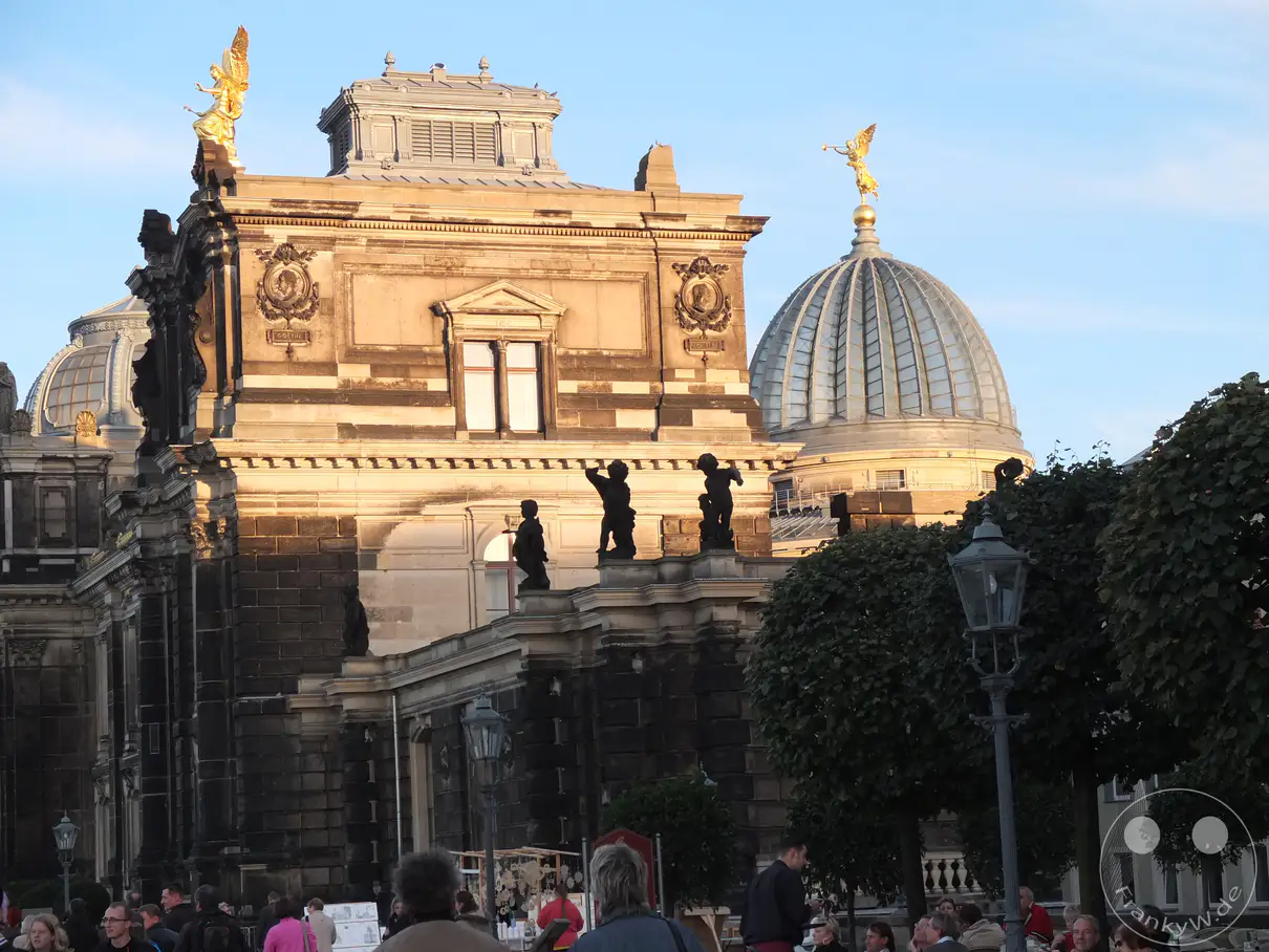 Die prächtige Architektur der Hochschule für Bildende Künste in Dresden wird von der goldenen Zitronenpresse auf der Kuppel und den Engelsfiguren überstrahlt. Die Szene ist in das warme Licht der Abendsonne getaucht, was eine friedliche und beeindruckende Atmosphäre schafft. Umgeben von Bäumen und belebten Plätzen, spiegelt das Bild die kulturelle Bedeutung und die historische Schönheit der Stadt wider.
