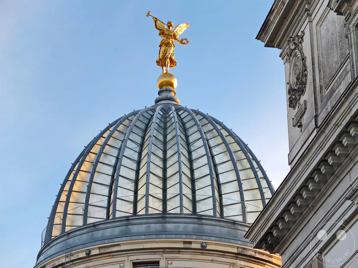 Die ber�hmte "Zitronenpresse", die eigentlich der Kuppel der Semperoper nachempfunden ist, wird von einer goldenen Engelsfigur bekr�nt. Die strahlende Sonne wirft ein warmes Licht auf die architektonischen Details des Geb�udes in Dresden, Deutschland. Der blaue Himmel bildet einen klaren Kontrast zum goldenen Engel und der Glas-Metall-Konstruktion der Kuppel und erzeugt eine majest�tische Atmosph�re.