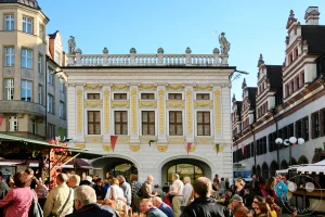 Die prachtvolle und reich verzierte Fassade der Alten Handelsbörse am Leipziger Naschmarkt bildet die beeindruckende Kulisse für ein belebtes Markttreiben im Stadtzentrum. Das im Jahr 1678 fertiggestellte barocke Prachtbauwerk besticht durch seine symmetrischen Fensterreihen, goldene Verzierungen und eine historische Balustrade mit Sandsteinstatuen auf dem Dach. Im Vordergrund drängen sich zahlreiche Besucher zwischen den herbstlich geschmückten Ständen der Leipziger Markttage, während rechts ein Teil des Alten Rathauses die historische Kulisse abrundet.