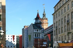 Die imposante Fassade des Steigenberger Grandhotel Handelshof erhebt sich am Rande des herbstlichen Markttreibens im Leipziger Stadtzentrum. Der monumentale ehemalige Messepalast besticht durch seine noble neoklassizistische Natursteinfassade mit gleichmäßigen Fensterreihen und eleganten Schriftzügen über den Fensterbögen. Im Vordergrund säumen die typischen hölzernen Stände und bunten Wimpel der Leipziger Markttage die Straße, während im Hintergrund der reich verzierte Turm des Alten Rathauses als historisches Wahrzeichen in den blauen Himmel ragt.