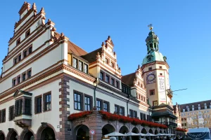 Das monumentale Alte Rathaus präsentiert sich an der Ostseite des großen Marktplatzes im historischen Stadtzentrum. Der weltberühmte Renaissancebau besticht durch seine markanten Zwerchhäuser, die langen Arkadengänge im Erdgeschoss und den asymmetrisch versetzten, prachtvollen Rathausturm mit der kupfernen Haube. Zahlreiche Passanten und Besucher beleben das gepflasterte Areal vor dem Gebäude, dessen Balkone im ersten Obergeschoss mit üppigem, rotem Blumenschmuck verziert sind und das historische Flair der Messestadt unterstreichen.
