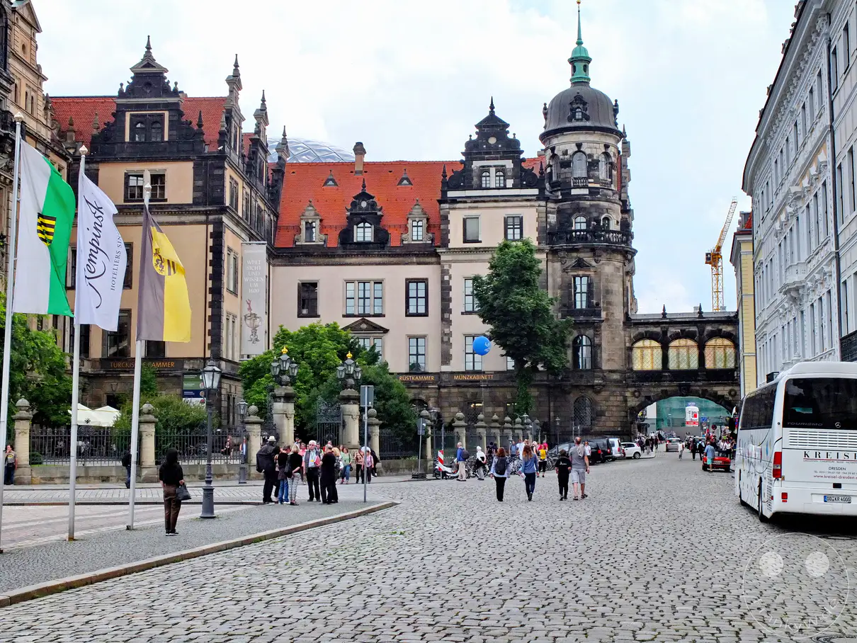 Das prächtige Dresdner Residenzschloss dominiert die Szenerie mit seiner kunstvollen Renaissance-Architektur und dem markanten Hausmannsturm auf der rechten Seite. Ein geschlossener Übergang verbindet das Schlossbauwerk über die Straße hinweg mit dem benachbarten Taschenbergpalais. Links wehen die sächsische Landesflagge sowie Werbefahnen im Wind, während mehrere Fußgänger die historische Pflasterstraße beleben. Am rechten Bildrand wartet ein weißer Reisebus auf dem weitläufigen Altstadtareal unter einem hellen Himmel.