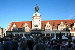 Die weitläufige Front des Alten Rathauses erstreckt sich majestätisch über die Ostseite des Leipziger Marktplatzes. Im Zentrum der Symmetrie ragt der prächtige, barock gedeckte Rathausturm empor, der von den charakteristischen, verzierten Zwerchhäusern des gewaltigen Renaissance-Daches flankiert wird. Im Vordergrund beleben zahlreiche Besucher das festliche Geschehen der Leipziger Markttage zwischen weiß-blauen Festzelten, Sonnenschirmen und gemütlichen Biertischgarnituren unter einem klaren, blauen Himmel.