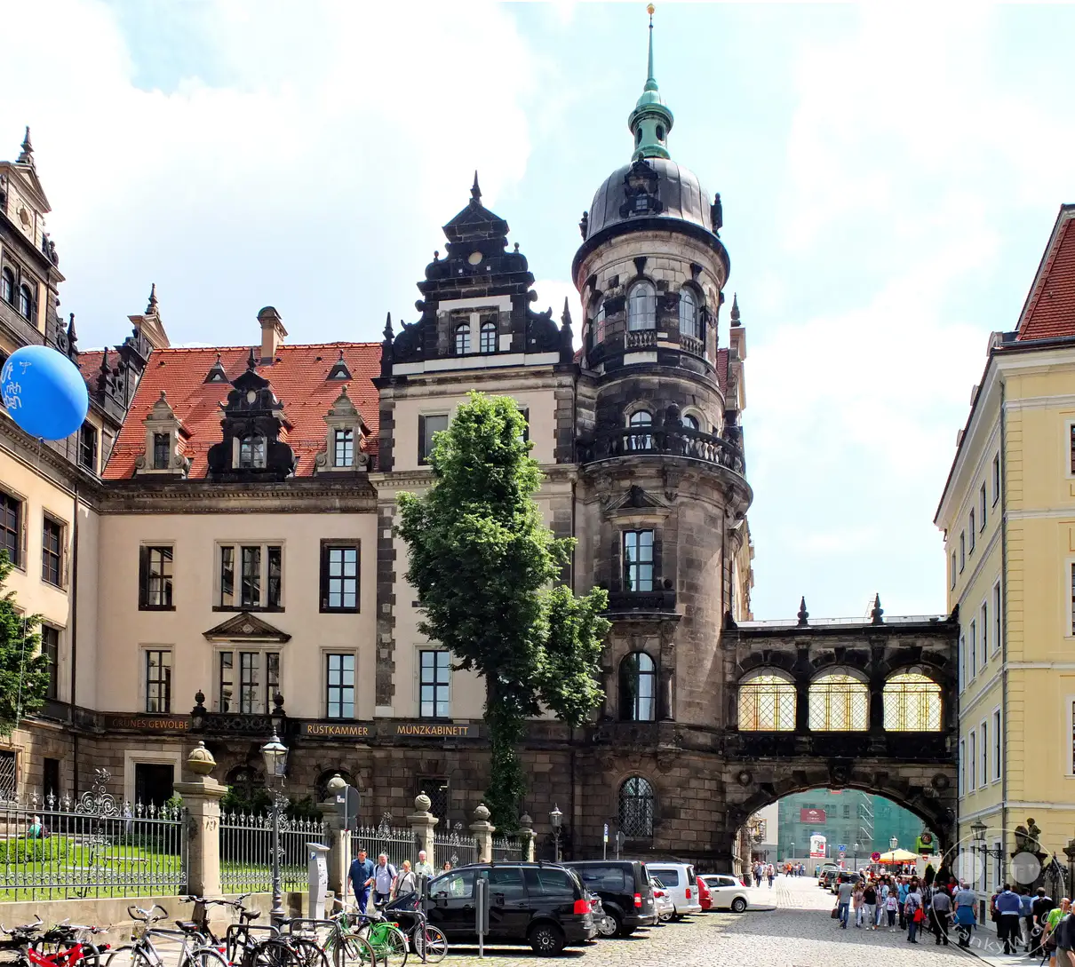 Das Dresdner Residenzschloss beeindruckt mit seinen reich verzierten Renaissance-Fassaden und dem markanten, barock gedeckelten Hausmannsturm auf der rechten Seite. Ein historischer Brückenübergang spannt sich elegant über die gepflasterte Straße im historischen Stadtkern. Vor dem verzierten Metallzaun des Schlossgeländes reihen sich zahlreiche Fahrräder an einem Fahrradständer aneinander, während vereinzelte Passanten das Areal passieren. Ein blauer Luftballon schwebt farbenfroh im Wind vor dem repräsentativen Eingangsbereich der Rüstkammer und des Münzkabinetts.
