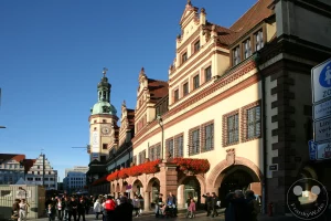 Die prachtvolle Rückfassade des Alten Rathauses präsentiert sich im warmen Sonnenlicht am historischen Leipziger Naschmarkt. Die detailreiche Architektur der Renaissance zeigt sich in den kunstvoll geschwungenen Zwerchhäusern, den rustikalen Arkadenbögen im Erdgeschoss und dem üppigen, roten Geranien-Blumenschmuck entlang der Balustrade. Zahlreiche Passanten flanieren über das historische Kopfsteinpflaster im Schatten des Gebäudes, während im Hintergrund der markante Rathausturm mit seiner barocken Haube in den klaren, blauen Himmel ragt.