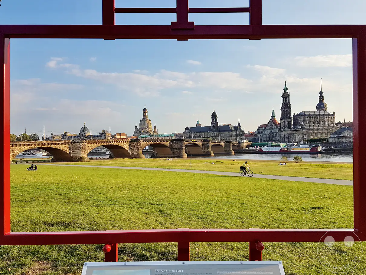 Ein sonniger Tag in Dresden, Deutschland, mit Blick auf die historische Augustusbrücke, die sich elegant über die Elbe spannt. Die Silhouette der Frauenkirche und anderer prachtvoller Gebäude bildet eine beeindruckende Kulisse, während eine Person mit dem Fahrrad eine grüne Uferpromenade entlangfährt. Die Szene strahlt eine friedliche und idyllische Atmosphäre aus, eingefangen durch einen dekorativen roten Rahmen.