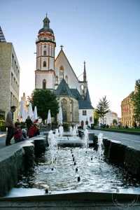 Die Thomaskirche in Leipzig bildet den imposanten Hintergrund für einen belebten Platz mit einem modernen Marktbrunnen im Vordergrund. Menschen genießen das sonnige Wetter und das urbane Treiben auf dem Augustusplatz. Die Szenerie vermittelt eine friedliche und lebendige Atmosphäre in der sächsischen Stadt.