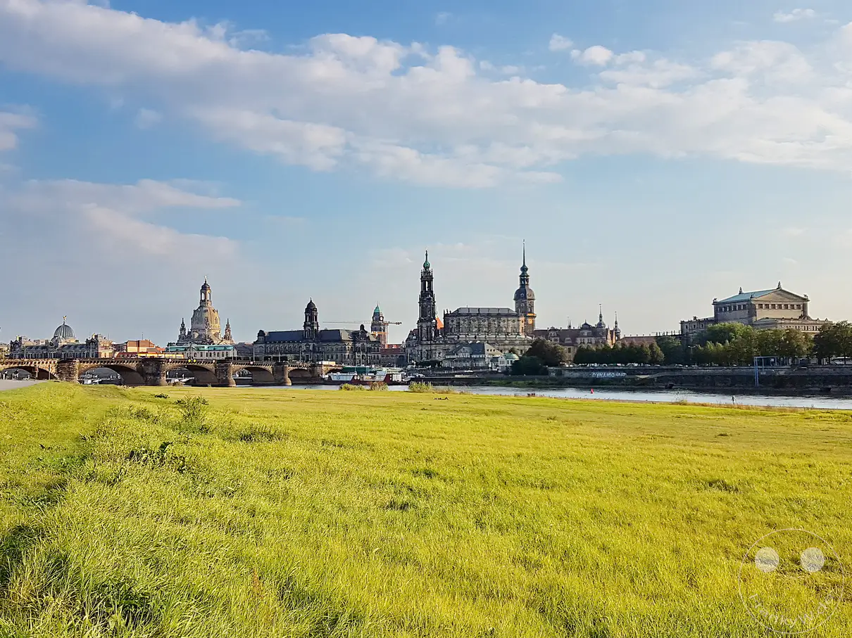 Ein malerischer Blick über die Elbe in Dresden offenbart die beeindruckende Architektur der Stadt, einschließlich der historischen Augustusbrücke und der Frauenkirche. Das sonnige Wetter mit leicht bewölktem Himmel verleiht der Szene eine friedliche und heitere Atmosphäre. Die üppig grüne Uferwiese lädt zum Verweilen ein und kontrastiert harmonisch mit den steinernen Monumenten am Horizont.