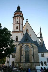Die Thomaskirche in Leipzig bildet den imposanten Hintergrund für einen belebten Platz mit einem modernen Marktbrunnen im Vordergrund. Menschen genießen das sonnige Wetter und das urbane Treiben auf dem Augustusplatz. Die Szenerie vermittelt eine friedliche und lebendige Atmosphäre in der sächsischen Stadt.