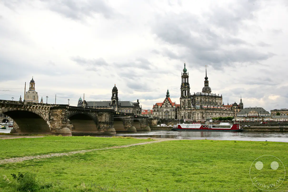 Die Elbfront von Dresden unter einem dramatischen Wolkenhimmel zeigt die imposante Augustusbrücke im Vordergrund. Im Hintergrund erheben sich historische Gebäude und Kirchtürme, während ein Ausflugsboot auf dem Fluss entlangfährt. Eine grüne Wiese mit einem steinernen Weg bildet den friedlichen Vordergrund dieser städtischen Ansicht.