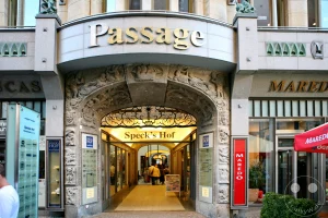 Die elegante Fassade eines historischen Passagengebäudes in Leipzig, Deutschland, fällt ins Auge. Ornamente und goldene Schriftzüge wie 'Passage' und 'Maredo' zieren die Eingänge. Der Durchgang 'Speck's Hof' lädt mit seinem warmen Licht und den Geschäften zu einem Besuch ein.