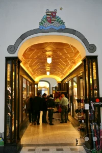 Ein prachtvoller Torbogen mit einem farbenfroh gestalteten, historischen Wappen markiert den Zugang zu einem der reich verzierten Gänge der Ladenpassage Specks Hof. Unter dem bogenförmigen Portal erstreckt sich ein elegant beleuchteter Tunnel, dessen kunstvoll strukturierte und goldglänzende Decke von warmen Leuchten erhellt wird. Zahlreiche Passanten flanieren durch den geschichtsträchtigen Gang, der von hölzernen Schaufensterfronten traditioneller Ladengeschäfte gesäumt ist und die exklusive Atmosphäre des sanierten Leipziger Messepalastes widerspiegelt.