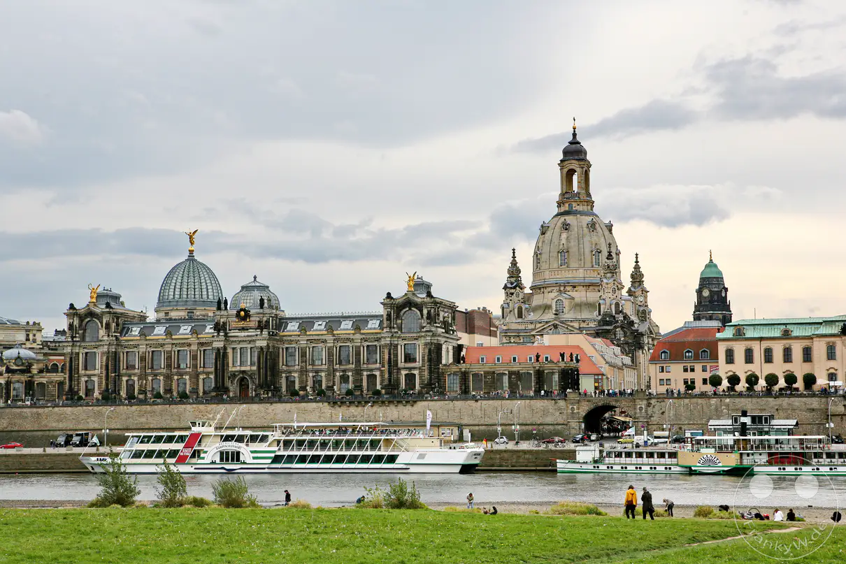 Die Silhouette von Dresden, mit den historischen Gebäuden der Altstadt, zeichnet sich am Ufer der Elbe ab. Sonnenschein und bewölkter Himmel schaffen eine weiche Lichtstimmung, während Ausflugsschiffe auf dem Fluss fahren. Menschen entspannen sich am grünen Uferstreifen, was die friedliche Atmosphäre der Stadt einfängt.