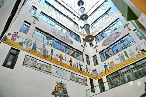 Der Blick nach oben offenbart die beeindruckende Innenarchitektur eines überdachten Lichthofes in der historischen Ladenpassage des Messepalastes Specks Hof. Die umlaufenden Etagenwände sind mit großflächigen, detailreichen Wandgemälden und bunten Mosaiken verziert, welche die Geschichte des Leipziger Messewesens und den Wandel des Handels künstlerisch darstellen. Eine moderne, trichterförmige Skulptur hängt zentral unter der gläsernen Dachkonstruktion, die viel natürliches Licht in den aufwendig sanierten und denkmalgeschützten Innenhof hineinlässt.